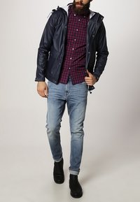 Marinblå vattentät jacka med vitfodrad huva, bärs över en röd och marinblå rutigt skjorta och ljusblå jeans; svarta anklar boots fulländar outfiten.