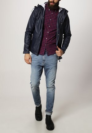 Marineblå vandtæt jakke med hvidforing på hætten, båret over en rød og marineblå ternet skjorte og lyseblå jeans; sorte ankelstøvler fuldender outfittet.