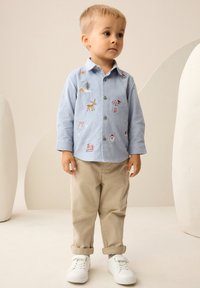 Hellblaues Hemd mit Knöpfen und bunten Stickereien, beigen Hosen und weißen Turnschuhen. Einfacher Kragen und hochgekrempelte Ärmel.