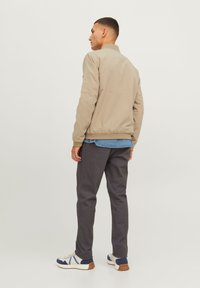 Veste bomber beige avec des poignets et un col côtelés, portée sur une chemise en denim, associée à un pantalon gris foncé et des baskets blanches décontractées.