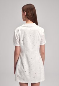 Valkoinen lyhythihainen mekko, jossa on teksturoitu kukkasilkkipisto- designs, kaulus ja vyötäröä korostava malli, joka levenee hieman helmassa.
