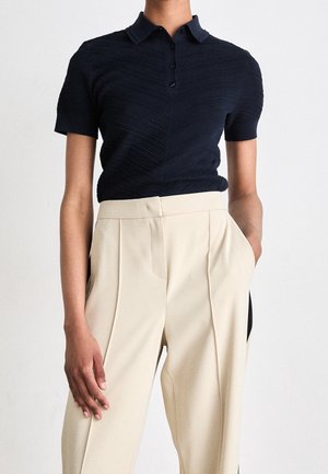 Personne portant un polo noir texturé à manches courtes rentré dans un pantalon plissé beige taille haute, les mains dans les poches.