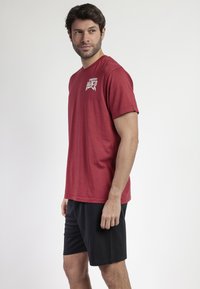 Camiseta de algodón roja con cuello redondo y mangas cortas, que presenta una impresión gráfica blanca en el lado izquierdo del pecho. Llevada con pantalones cortos negros.
