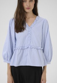 Bluza cu dungi bleu, decolteu în formă de V, detalii cu volane de-a lungul gâtului și tivului, și mâneci bufante. Materialul pare moale și texturat.