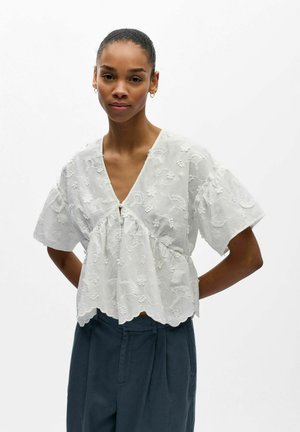 Frau trägt eine weiße, mit Blumen bestickte Bluse mit kurzen Ärmeln und gezacktem Saum sowie eine dunkle, hoch taillierte Hose, steht vor weißem Hintergrund.