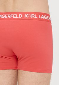 Boxer en coton rouge avec une ceinture blanche portant un logo. Le design présente une texture lisse et une coupe ajustée.