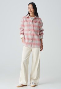 Roze en witte gestreepte flanellen shirtjack met twee borstzakken, knoopsluiting en witte accenten, gecombineerd met crèmekleurige wijde broek en sneakers.