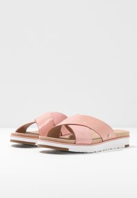Roze sandalen met een glanzend kruisbandontwerp, voorzien van een zachte suède-achtige textuur en een witte gestructureerde zool met lichtbruine accenten.