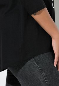 Černé bavlněné tričko s krátkými rukávy. Má zakřivený lem a volnější střih. Kombinováno s tmavě šedými džínami. Hladká textura.