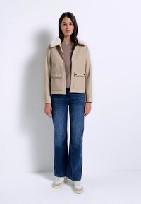 Frau steht und trägt eine beige Jacke mit weißem Kragen, taupefarbenes Oberteil, blaue Jeans mit weitem Bein und weiße Sneakers vor einem weißen Hintergrund.
