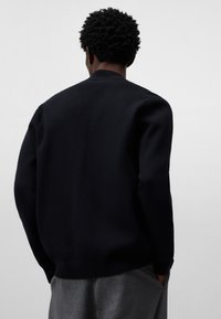 Svart stickad tröja med rundad hals och ribbade muddar, med en texturerad yta. Bärs med grå byxor, sedd bakifrån.