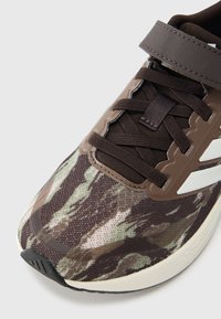 Καφέ και πράσινο αθλητικό παπούτσι με σχέδια, με υφασμένο επάνω μέρος, μαύρα κορδόνια και λευκή διακοσμητική λεπτομέρεια με λογότυπο στο πλάι.