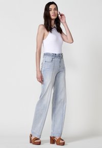 Jean large bleu clair avec un détail de ceinture tressée, associé à un débardeur blanc et des sandales compensées marron.