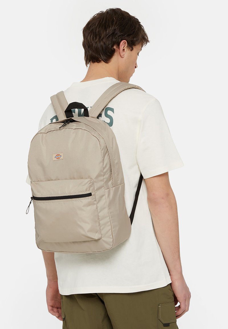 Mochila de tecido bege com forma arredondada, fecho preto, bolso frontal, alças ajustáveis e um pequeno logótipo no painel frontal superior.