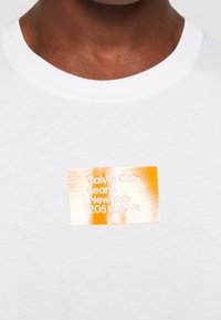 Bílý bavlněný tričko s obdélníkovou oranžovou a stříbrnou reflexní nášivkou s textem "Calvin Klein Jeans New York 205 W39 St.".
