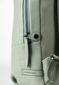 Hellgrüner Leder-Rucksack mit einem silbernen Reißverschluss, einer Stoffauskleidung und einem gestickten Logo. Verfügt über eine kleine Seitentasche und ein metallisches Akzent.
