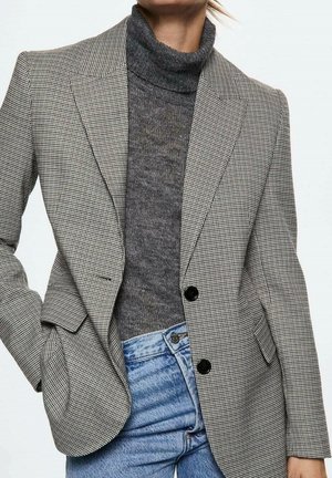 Kvinde iført en grå hundetandsblazer over en mørkegrå rullekravetrøje og lyseblå jeans, hånd i blazerlommen.