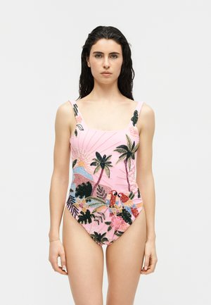 Γυναίκα με σκούρα μαλλιά φοράει ροζ μαγιό διακοσμημένο με κεντημένη τροπική σκηνή που περιλαμβάνει φοίνικες, λουλούδια και πολύχρωμους παπαγάλους.