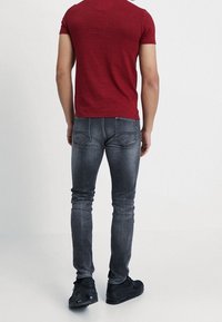 Hombre vestido con jeans negros ajustados y desgastados, una camisa roja de manga corta y zapatillas negras, de pie con la espalda hacia la cámara.