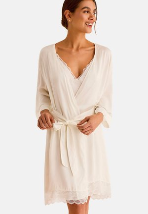 Femme souriante, portant un peignoir crème orné de dentelle, noué à la taille, avec des manches trois-quarts et un caraco à col en V en dessous.