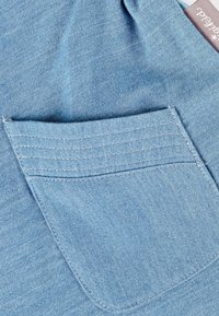 Helles blaues Denimgewebe mit einer Vordertasche, die über eine detaillierte Nahtführung und eine glatte Textur verfügt, zeigt sein lässiges Design und seinen praktischen Nutzen.