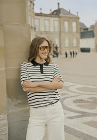 Gestreepte polo met zwarte kraag en korte mouwen; gecombineerd met witte broek. Ronde gele zonnebril voegt een levendige toets toe. Achtergrond van een stenen muur.