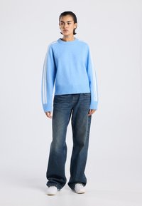 Pull en tricot bleu clair avec ourlet côtelé, arborant des rayures blanches le long des manches, associé à un jean en denim taille haute et ample.
