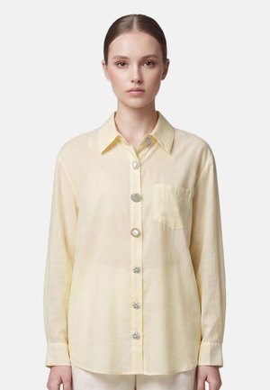 Vrouw draagt een lichtgele blouse met lange mouwen, unieke decoratieve knopen en een borstzak, staand tegen een effen achtergrond.