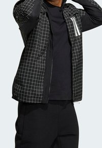 Mustat vetoketjullinen takki, jossa on ruudukkomalli hihoissa, musta runko ja valkoinen korostuslogo rinnassa.