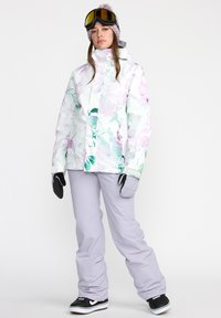 Dames sneeuwjack met bloemenpatroon in wit, paars en groen. Gecombineerd met lichtgrijze sneeuwbroek en zwarte sneakers, met zwarte handschoenen.