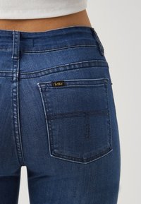 Jean en denim bleu foncé avec une coupe ajustée, présentant une poche arrière distincte et une étiquette logo "Lois" près du haut de la poche.