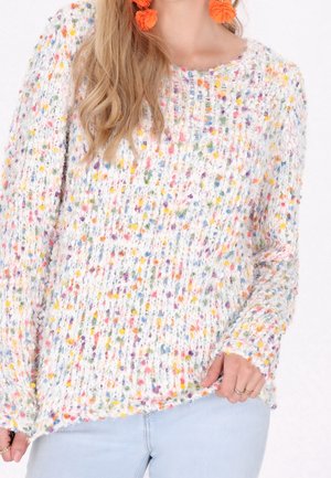 Femme portant un pull en tricot blanc avec des taches multicolores, un jean bleu clair, des boucles d'oreilles florales orange et des bagues aux doigts.