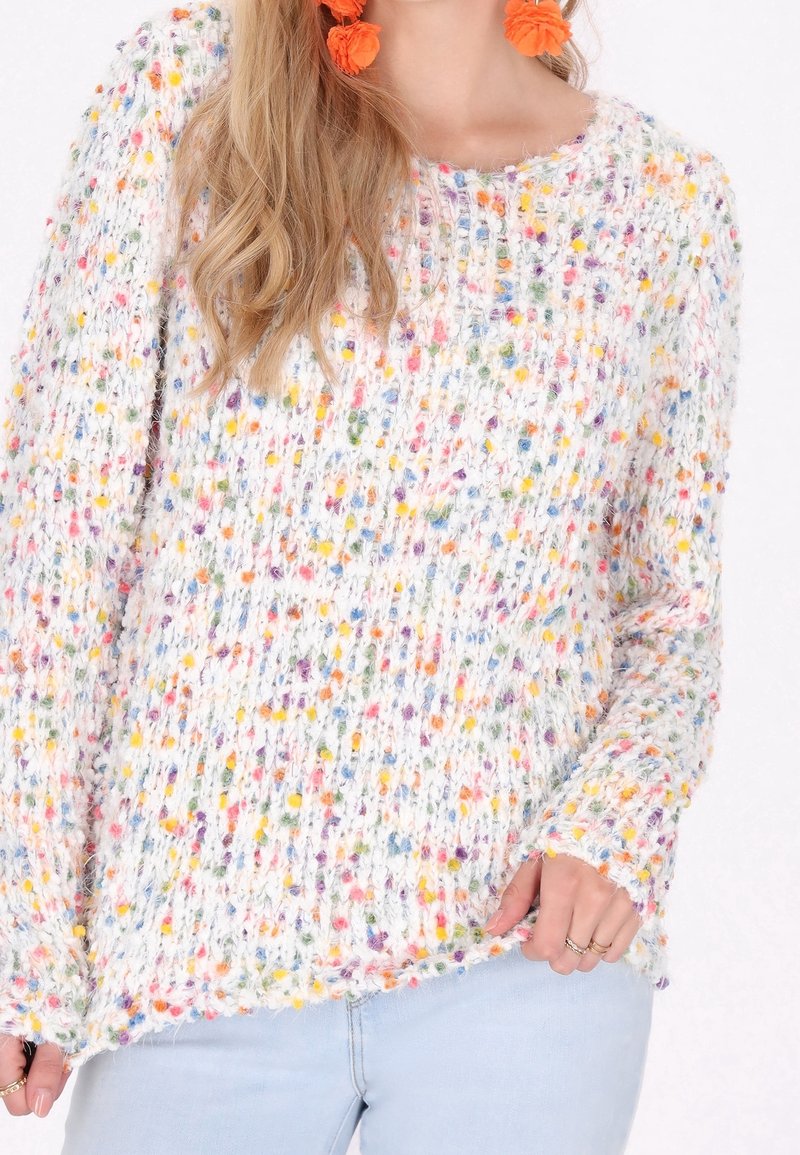 Femme portant un pull en tricot blanc avec des taches multicolores, un jean bleu clair, des boucles d'oreilles florales orange et des bagues aux doigts.