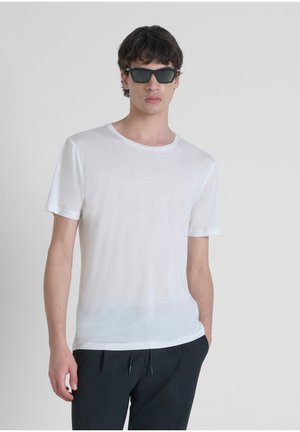 T-shirt bianca a maniche corte con scollatura rotonda e tessuto leggero, leggermente trasparente, abbinata a occhiali da sole neri e pantaloni scuri.