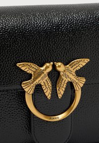 Μαύρη τσάντα από υφή δέρματος με χρυσό κυκλικό κούμπωμα, διακοσμημένο με δύο λεπτομερείς φιγούρες πουλιών. Ονομασία της μάρκας "PINKO" είναι εμφανής.