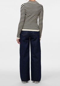 Femme portant un haut à manches longues rayé noir et blanc et un jean large foncé, debout de dos sur un fond uni.