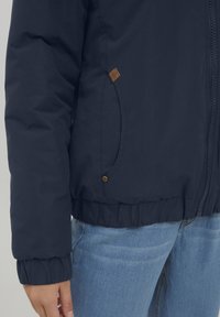 Giacca puffer blu navy con polsini a coste e chiusura con zip, dotata di una tasca laterale e di un'etichetta in pelle marrone sul fondo.