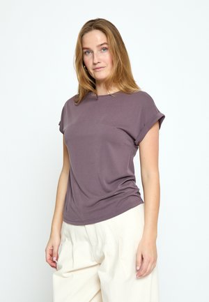 Ung kvinde med langt blondt hår iført en mauve kortærmet top og løse hvide bukser mod en ensfarvet hvid baggrund.