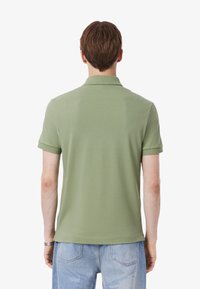 Modello che indossa una polo a maniche corte, di colore verde oliva, girato di spalle, abbinata a jeans di denim azzurri. Sfondo neutro.
