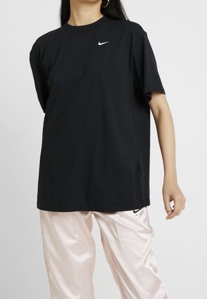 Osoba ubrana w czarną koszulkę Nike z białym logo oraz jasnoróżowe satynowe spodnie na jednolitym tle.