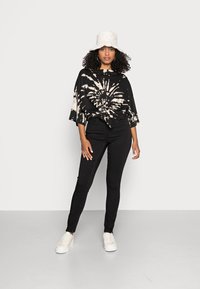 Top oversized com padrão tie-dye preto e padrões em bege claro, combinado com jeans skinny pretos de cintura alta e ténis brancos. Chapéu de balde branco.