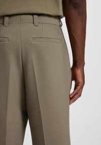 Close-up of a person’s lower back and hand, wearing beige tailored trousers and a matching tucked-in shirt.
