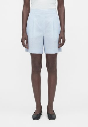 Nő világoskék, texturált, magas derekú, elöl redőzött rövidnadrágot, fehér felsőt és fekete balettcipőt visel, egy egyszínű háttér előtt áll.