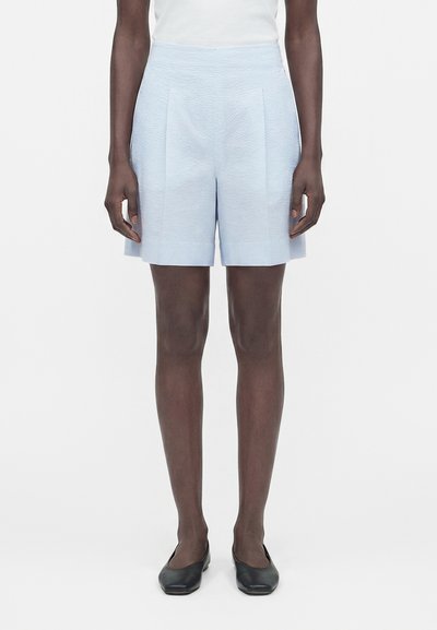 Femme portant un short taille haute texturé bleu clair avec des plis sur le devant, un haut blanc et des ballerines noires, debout devant un fond uni.