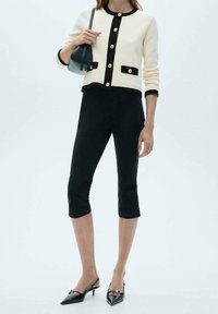 Žena v bílém kardiganu s černými lemováními a zlatými knoflíky, černých zkrácených kalhotách, černých lodičkách na podpatku a dr�žící černou kabelku.