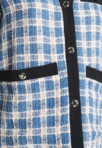 Top in tessuto intrecciato blu e bianco con motivo, caratterizzato da una pattina frontale nera, bottoni grandi e due tasche con rifinitura nera uniforme.