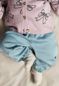 Lys rosa cardigan med svarte sløyfemønstre, gullknapper; blå bukser i denimstil med elastiske mansjetter; hvite sokker synlige.