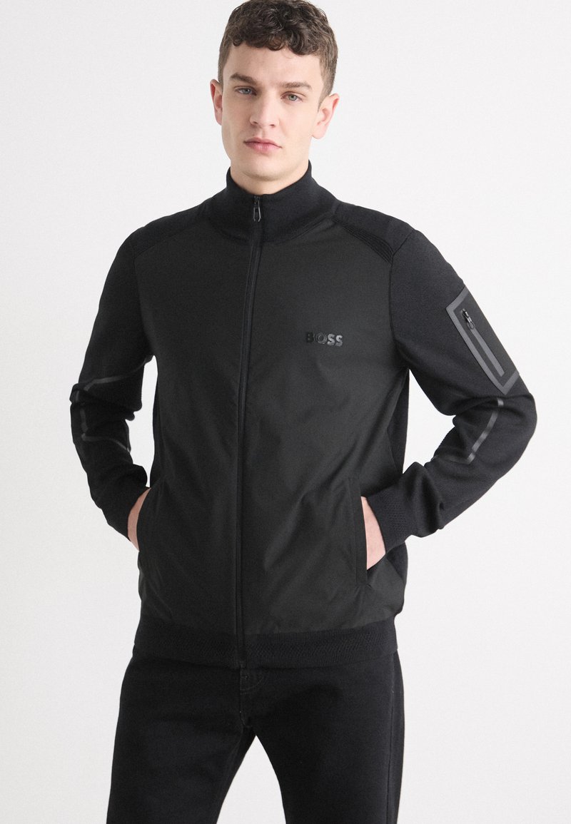 Black zip-up jacket with a high collar, featuring a subtle logo, textured accents, and a zip pocket on the sleeve.