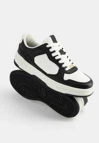 Black and white sneakers with a leather upper, perforated toe, flat white laces, and a textured black rubber sole. Gold accent on the tongue.