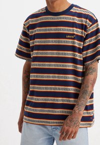 Krátký rukáv námořnické pruhované tričko s horizontálními vzory v béžové a červené barvě, s malým žlutým nápisem "Levi's" na hrudi.